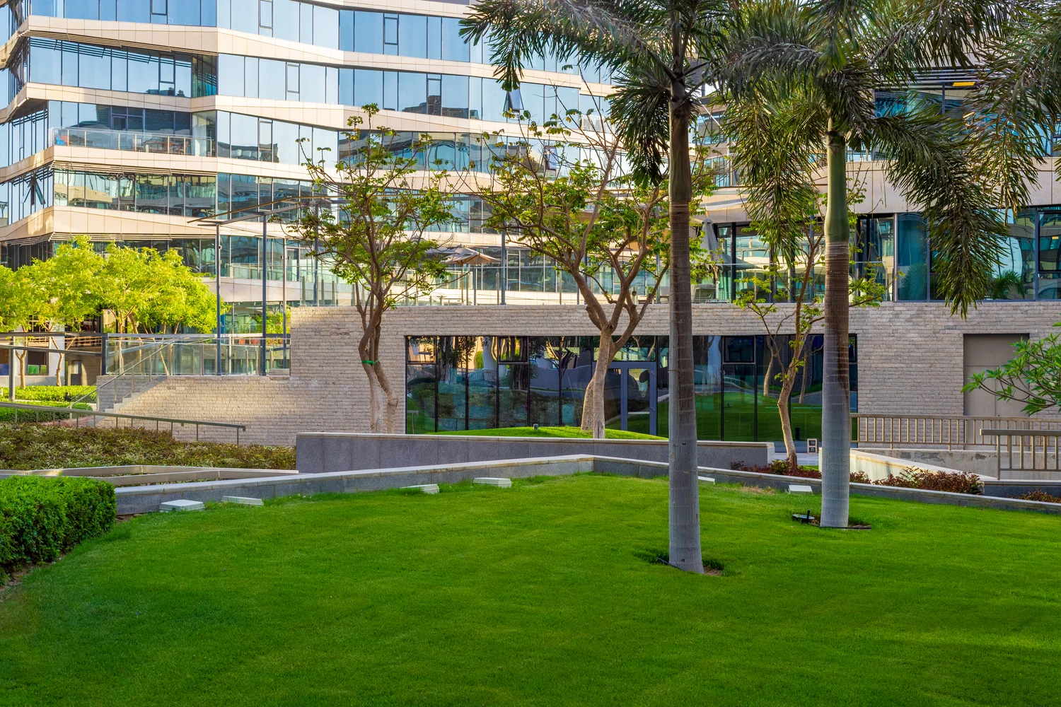 Modern office building with large glass windows, surrounded by a neatly landscaped garden featuring grass, trees, and pathways.