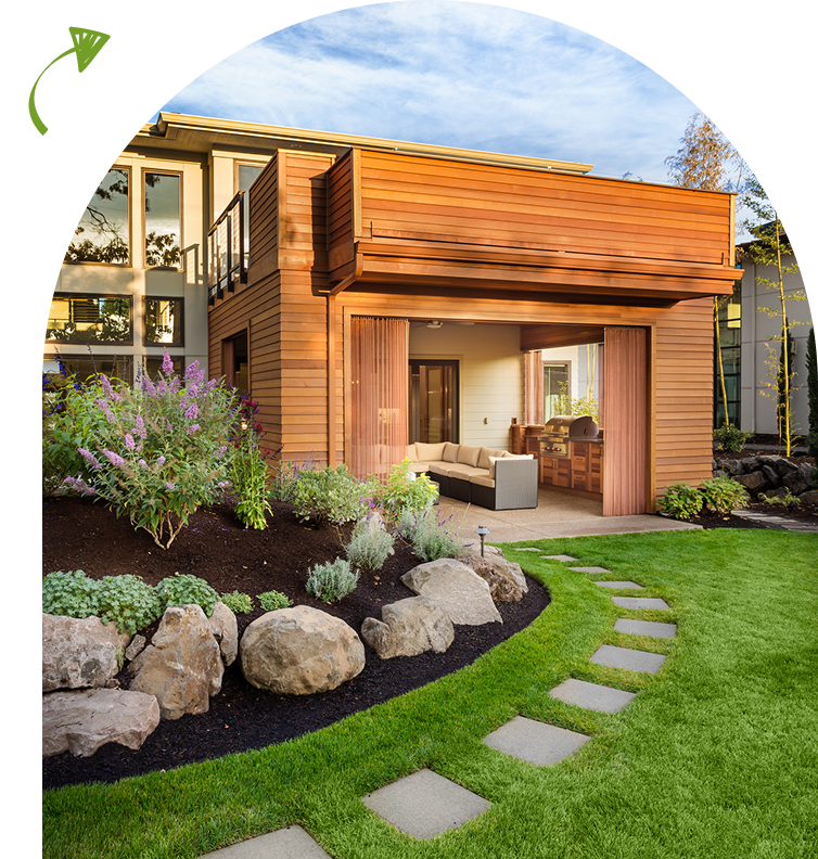 Modern wooden house with a large deck, surrounded by a landscaped garden with stone path and flowering plants. Patio furniture visible inside.