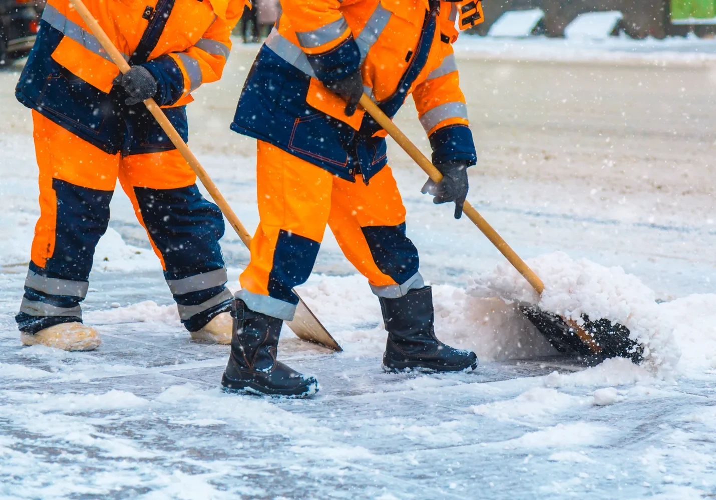 Two workers in orange reflective gear shovel snow on a street during a snowfall.
