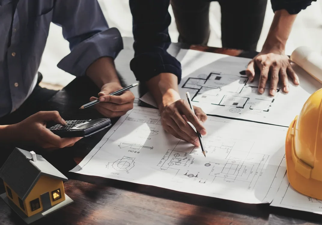 Two people reviewing architectural blueprints on a table with a smartphone, calculator, model house, and yellow hard hat nearby.